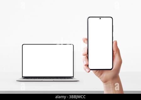 Smartphone in hand with blank screen and laptop in background on desk, both ready for UI or branding mockup Stock Photo