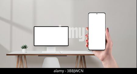 Phone in hand and computer monitor in background, both with blank white screens, ideal for app and web design mockup Stock Photo