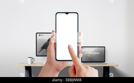 Smartphone mockup in female hands with finger touching white screen, office background with laptop and monitor Stock Photo
