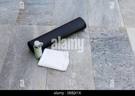 Rolled black exercise mat lying diagonally on grey patio with white towel and green water bottle Stock Photo