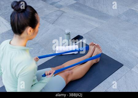 Pulling Asian woman in mint activewear using resistance band on mat on patio, with water bottle Stock Photo