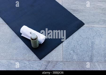 Yoga mat is lying diagonally on grey patio with rolled towel, green water bottle, copy space Stock Photo