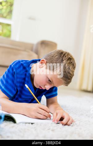 Boy lying on white carpet in bright living room, writing in open notebook with yellow pencil Stock Photo