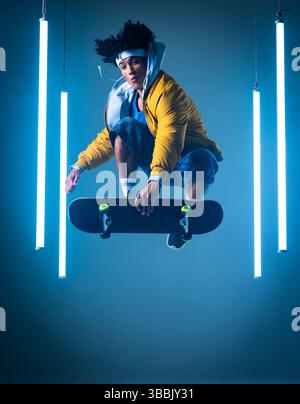 Jumping African American man in yellow jacket holding skateboard in blue studio, with neon tubes Stock Photo