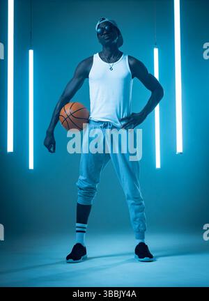 Posing African American man wearing athletic attire in blue studio, with basketball and light tubes Stock Photo