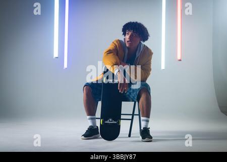Sitting man wearing mustard yellow jacket posing in studio, holding black skateboard with LED tubes Stock Photo