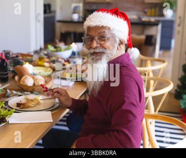 Senior man wearing Santa hat sitting at dining table at home, holding fork with red berries Stock Photo
