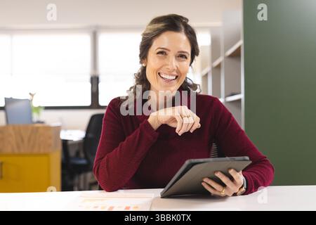 Businesswoman sitting at desk in bright modern office, holding tablet and displaying bar charts Stock Photo