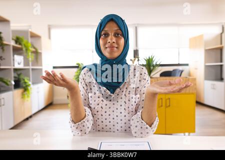 Woman shrugging at desk in modern office, with papers, potted plants and yellow cabinet Stock Photo
