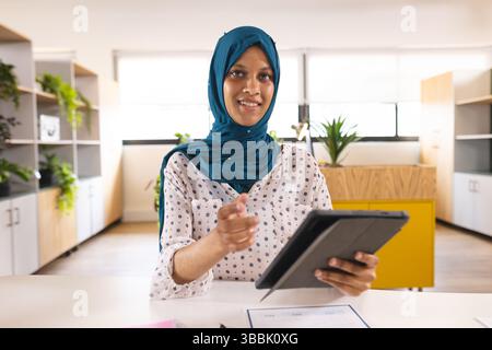 Woman wearing teal hijab extending hand and holding tablet in modern office, with papers Stock Photo