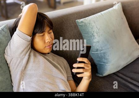 Young Asian man reclining on gray couch at home with teal pillows, using smartphone, copy space Stock Photo