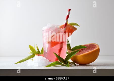 Refreshing grapefruit drink with ice in a glass cup decorated with a straw and a slice of fruit on a white table with fruit, ice and leaves with a whi Stock Photo