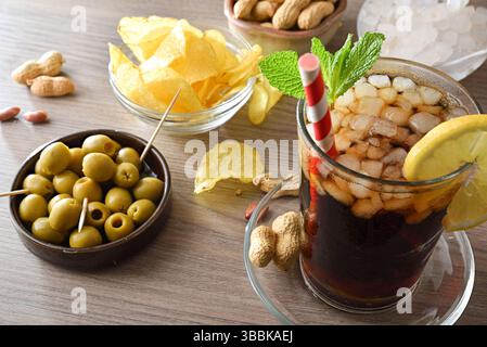 Refreshing cola drink and snack containers on a wooden table with olives, potatoes, and peanuts. Elevated view. Stock Photo
