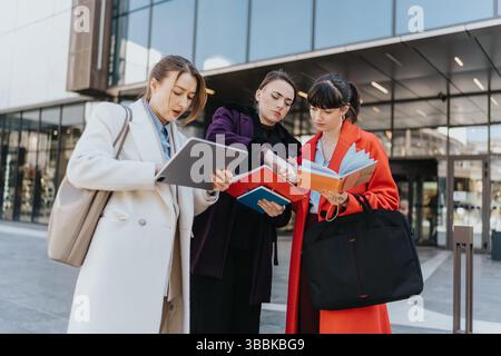 Three professional women collaborating outside a modern building while reviewing documents and a tablet Stock Photo