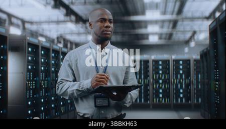 Big Data Center with Server Racks: Professional African American Specialist Using Tablet Computer, Activating Servers, Standing In Warehouse. Information Technology and Cyber Security. Medium Shot. Stock Photo