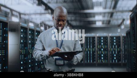 Big Data Center with Server Racks: Professional African American Specialist Using Tablet Computer, Activating Servers, Standing In Warehouse. Information Technology and Cyber Security. Medium Shot. Stock Photo