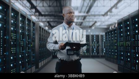 Big Data Center with Server Racks: Professional African American Specialist Using Tablet Computer, Activating Servers, Standing In Warehouse. Information Technology and Cyber Security. Medium Shot. Stock Photo