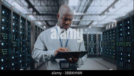 Big Data Center with Server Racks: Professional African American Specialist Using Tablet Computer, Activating Servers, Standing In Warehouse. Information Technology and Cyber Security. Medium Shot. Stock Photo