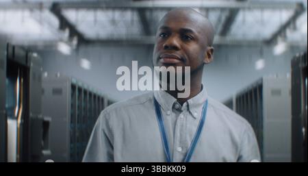 Professional African American Specialist, Administrator Walking in Big Data Center with Server Racks, Smiling, Looking at Camera. Internet Servers, Information Technology and Cyber Security Concept. Stock Photo