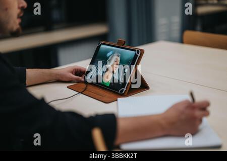 Young man participating in an online video call while taking notes on a notepad at a table Stock Photo