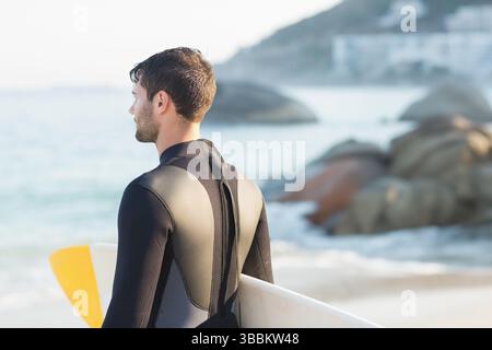 White surfboard resting on sandy shoreline facing calm ocean with rocky boulders under clear sky Stock Photo