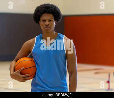 Young African American man standing on gymnasium court, holding orange basketball under arm Stock Photo