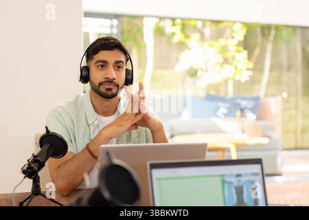 Young Indian man sitting at desk in home office, wearing headphones and using laptop and microphone Stock Photo