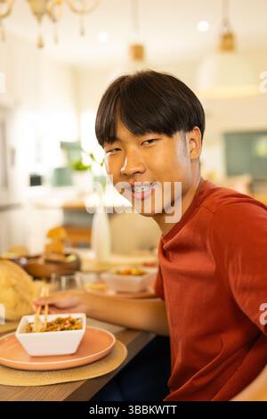 Young Asian man eating noodles from square bowl with chopsticks at table in modern dining room Stock Photo