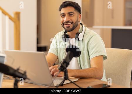 Indian man sitting at desk in home studio, with laptop, microphones, headphones and smartphone Stock Photo
