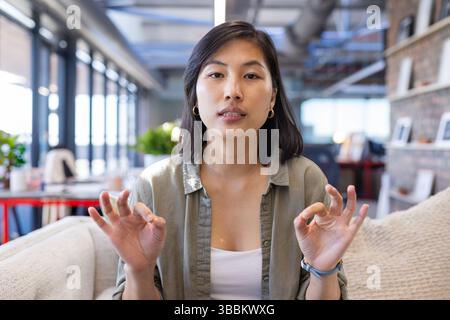 Young Asian woman making OK gesture while sitting on couch in modern office, showing confidence Stock Photo