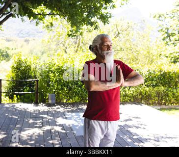 Senior man practicing standing yoga pose on wooden deck with yoga mat and planter, copy space Stock Photo