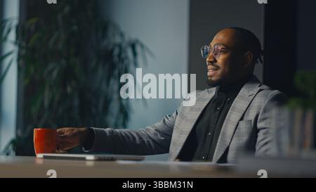 Confident entrepreneur or consultant sitting at desk in office, reaching for coffee while engaged in thoughtful conversation. Concept of leadership Stock Photo