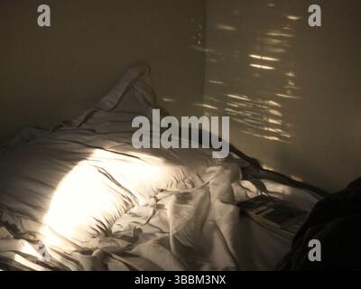 Morning sunlight casting shadows on an unmade bed with a pillow and book Stock Photo