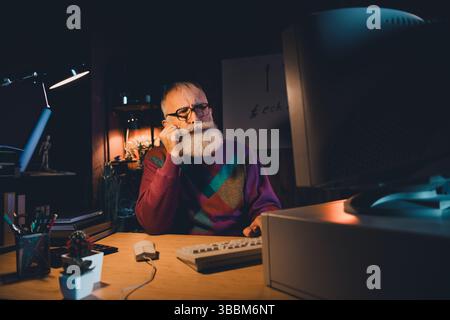 Elderly programmer working late at home office desk, wearing colorful sweater, focused on computer coding development Stock Photo