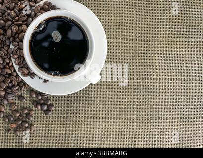 A steaming cup of black coffee sits on white saucer, surrounded by scattered coffee beans on textured burlap background, creating warm and inviting at Stock Photo