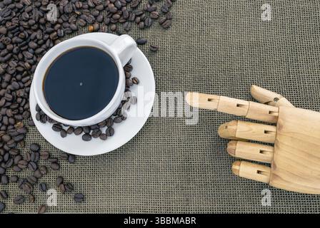 A white coffee cup filled with black coffee sits on saucer surrounded by coffee beans on textured surface, with wooden hand pointing towards it Stock Photo