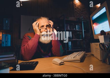 Elderly man working on computer in a home office setting, displaying stress in a casual atmosphere with warm evening light Stock Photo