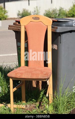 A wooden chair with a brown cushion is sitting on the grass next to a trash can Stock Photo