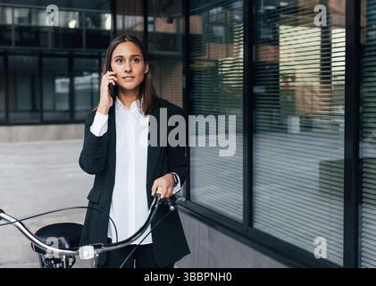 Confident female office worker with a bicycle talking on mobile phone outdoors Confident female office worker with a bicycle talking on mobile phone o Stock Photo