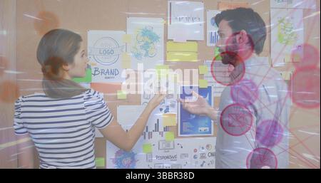 Collaborating team analyzing brand sketches on cork bulletin board in office, with sticky notes Stock Photo
