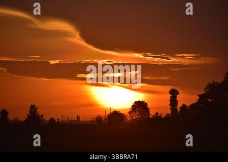 A glowing sun sets behind dark tree silhouettes, casting a fiery orange hue across the sky. This peaceful sunset scene captures the serene transition Stock Photo