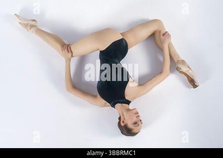 A young ballerina in a dark bodysuit lying on the floor shows stretching and eversion of her arms Stock Photo