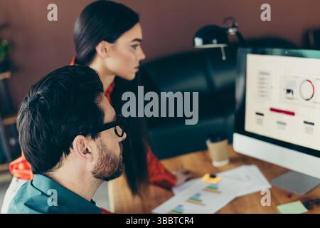 Focused coworkers collaborating on business analytics in a modern workspace, analyzing data graphs and charts on a desktop computer Stock Photo