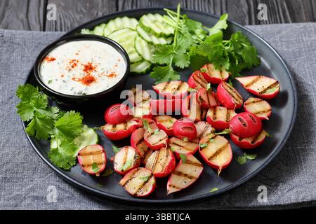 Grilled radishes with cucumber slices and sour cream sauce served on a dark grey plate on a black wooden table, horizontal view, real photo, not AI ge Stock Photo