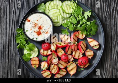 Grilled radishes with cucumber slices and sour cream sauce served on a dark grey plate on a black wooden table, top view, flat lay, close-up, real pho Stock Photo