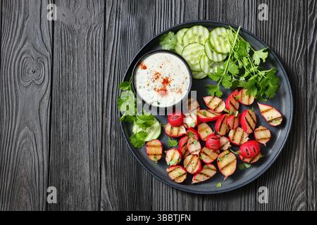 Grilled radishes with cucumber slices and sour cream sauce served on a dark grey plate on a black wooden table, top view, flat lay, free space, real p Stock Photo
