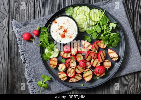 Grilled radishes with cucumber slices and sour cream sauce served on a dark grey plate on a black wooden table, top view, flat lay, real photo, not AI Stock Photo