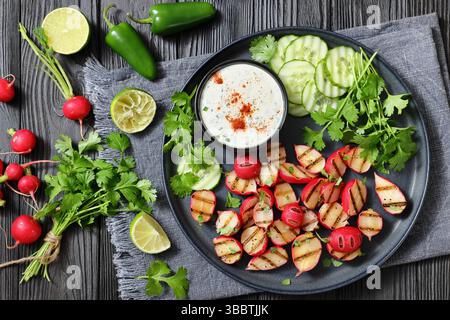 Grilled radishes with cucumber slices and sour cream sauce served on a dark grey plate on a black wooden table with fresh cilantro, jalapeno pepper, l Stock Photo