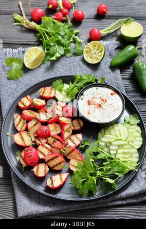 Grilled radishes with cucumber slices and sour cream sauce served on a dark grey plate on a black wooden table with fresh cilantro, jalapeno pepper, l Stock Photo