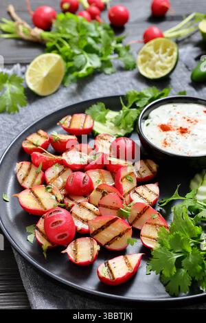 Grilled radishes with cucumber slices and sour cream sauce served on a dark grey plate on a black wooden table with fresh cilantro, jalapeno pepper, l Stock Photo